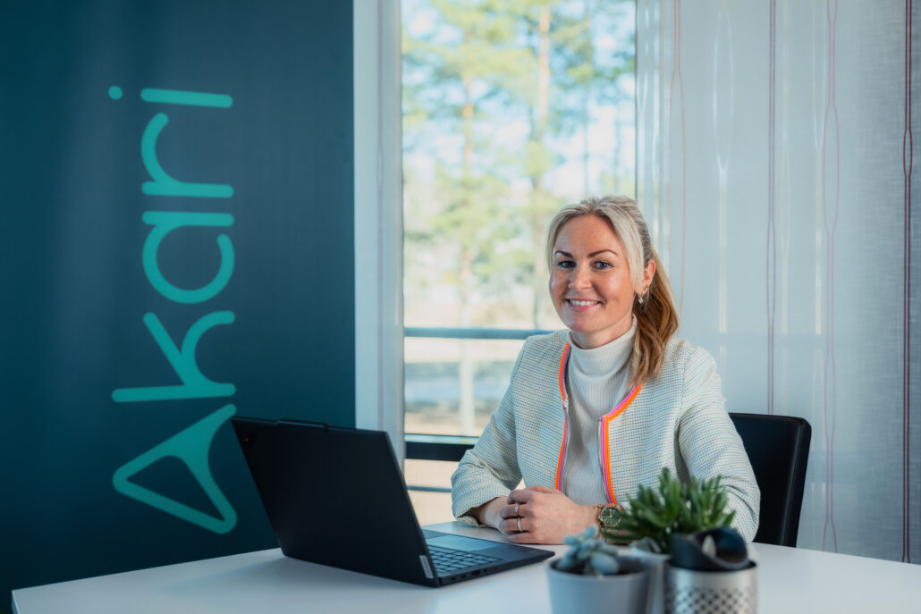 Christina i Akari sitter på Hønefoss-kontoret, ved pcen, foran en rollup med Akari-logo på