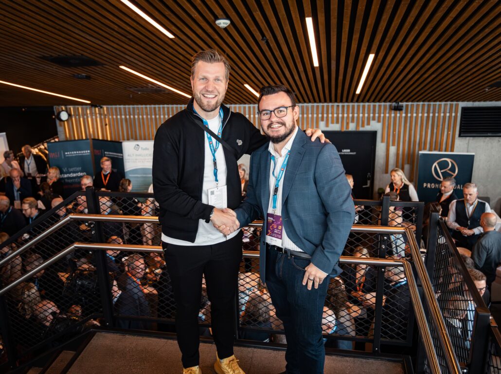Nicolay Bell smiler og håndhilser med Christian Christian Aarbogh Bjerke foran messeområdet under Ringerikekonferansen 2025. Begge har deltakerbånd og navneskilt, og i bakgrunnen er det mange mennesker i samtaler mellom stands og utstillere.
