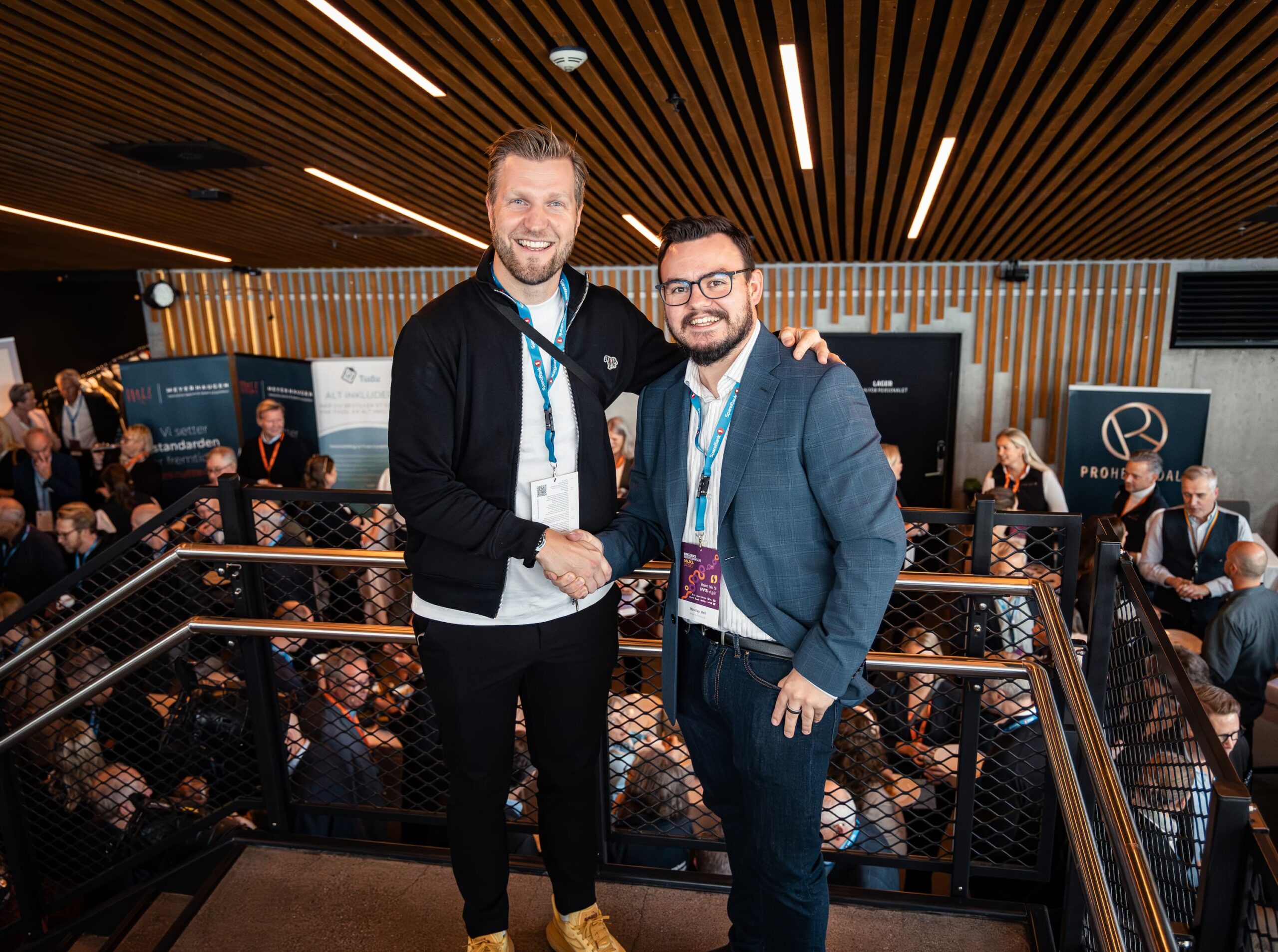 Nicolay Bell smiler og håndhilser med Christian Christian Aarbogh Bjerke foran messeområdet under Ringerikekonferansen 2025. Begge har deltakerbånd og navneskilt, og i bakgrunnen er det mange mennesker i samtaler mellom stands og utstillere.