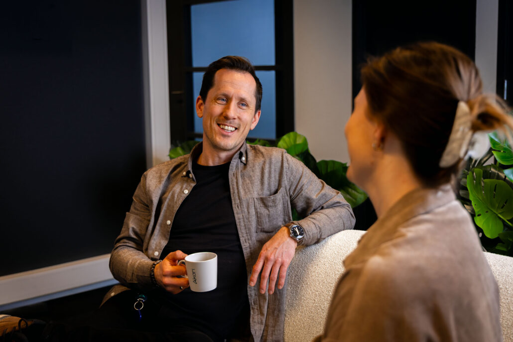 Geir smiler mot sin kollega Pia mens han holder en kaffekopp i hånden