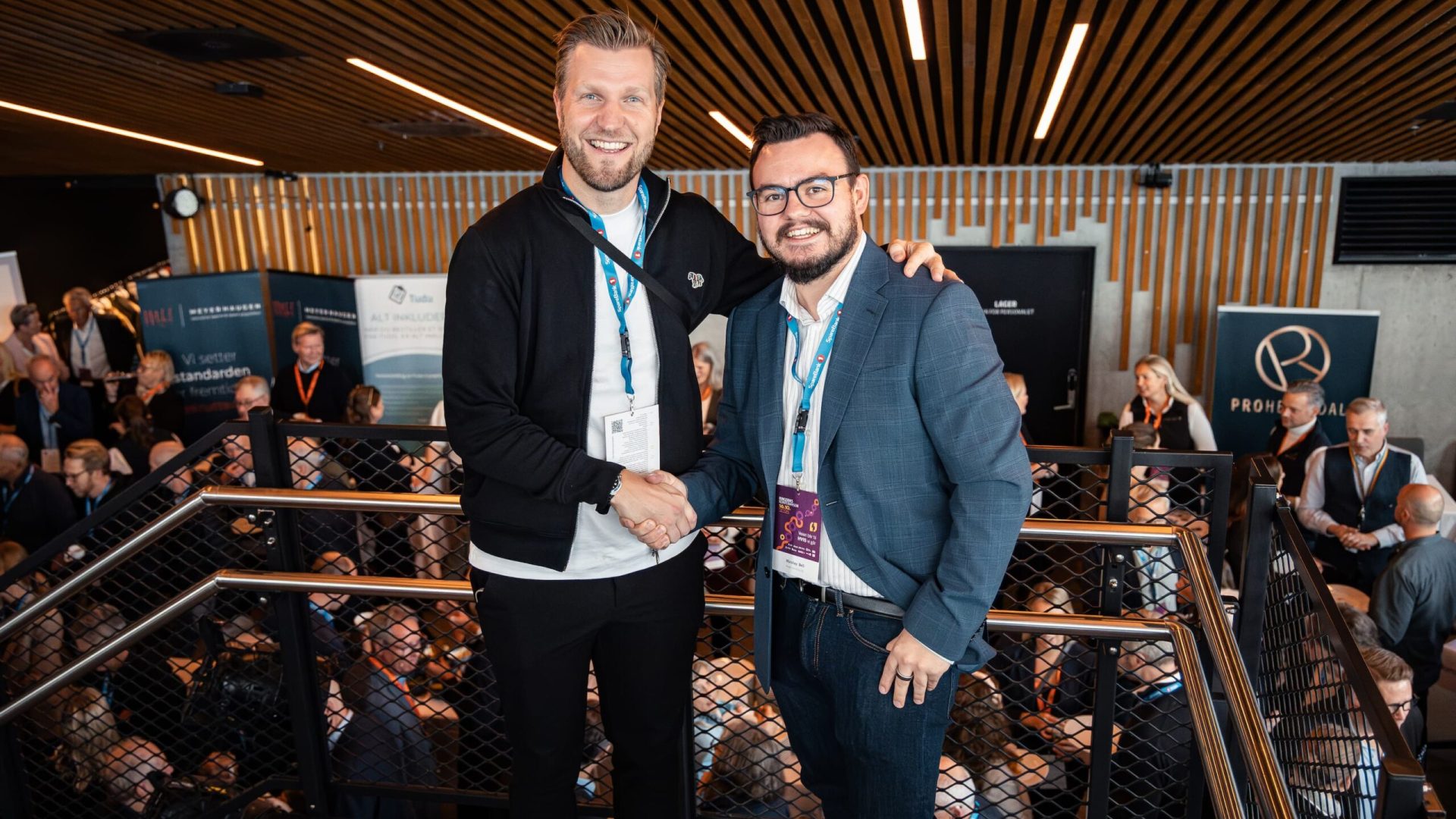 Nicolay Bell smiler og håndhilser med Christian Christian Aarbogh Bjerke foran messeområdet under Ringerikekonferansen 2025. Begge har deltakerbånd og navneskilt, og i bakgrunnen er det mange mennesker i samtaler mellom stands og utstillere.