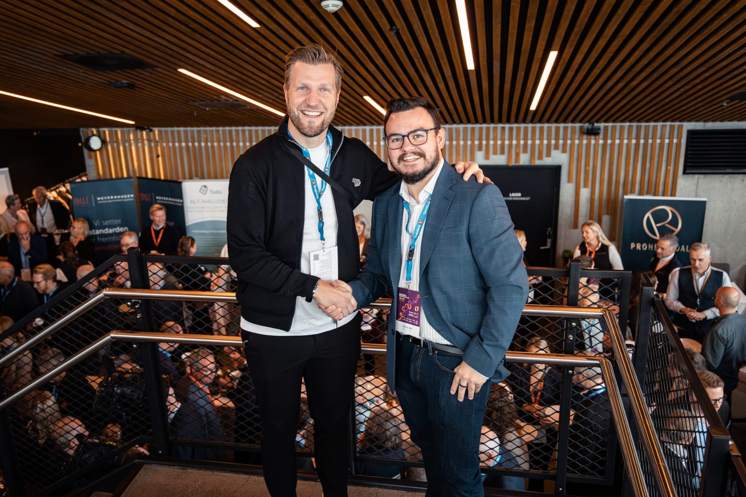 Nicolay Bell smiler og håndhilser med Christian Christian Aarbogh Bjerke foran messeområdet under Ringerikekonferansen 2025. Begge har deltakerbånd og navneskilt, og i bakgrunnen er det mange mennesker i samtaler mellom stands og utstillere.